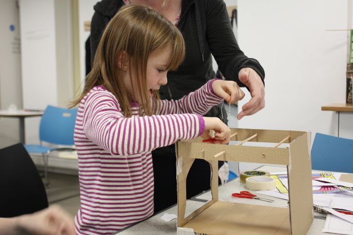 A child builds a small cardboard structure at a table while an adult assists with the craft materials.