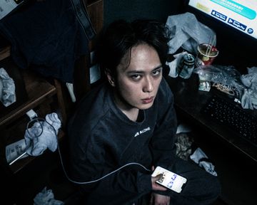 A person sits in a cluttered room holding a smartphone connected to a charging cable, surrounded by discarded packaging, clothing, and an illuminated computer screen on a desk.