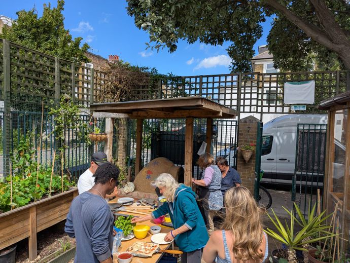 People gather in a community garden, preparing food together beside an outdoor clay oven.