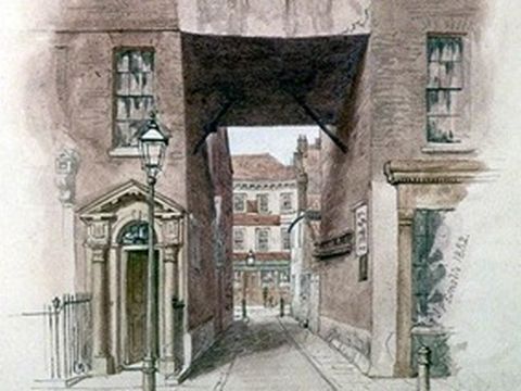 A narrow cobbled street between old multi-storey brick buildings with an archway and a lamp post in the foreground.