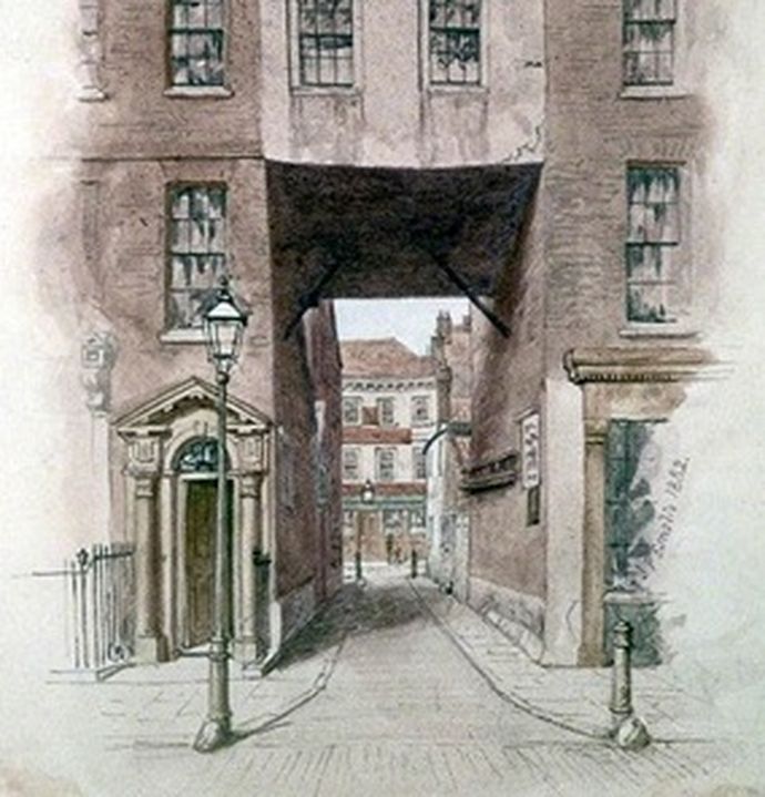 A narrow cobbled street between old multi-storey brick buildings with an archway and a lamp post in the foreground.
