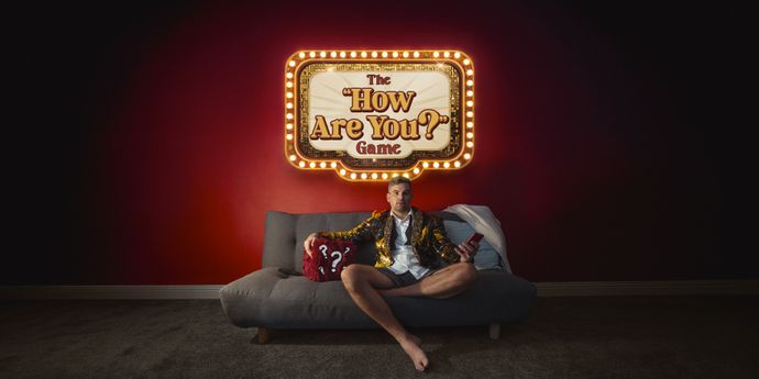 Man sitting on a couch holding a question mark cushion under a lit sign that reads "The How Are You? Game" against a red wall.