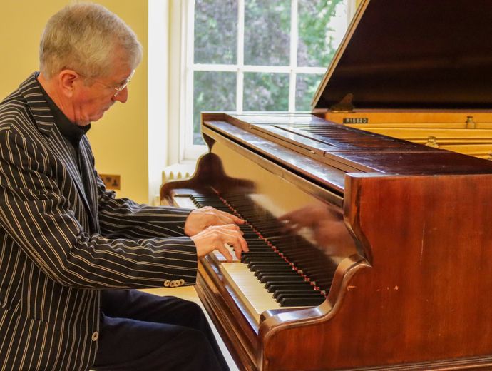 A person plays a polished grand piano indoors, with sunlight streaming through a window behind the instrument.
