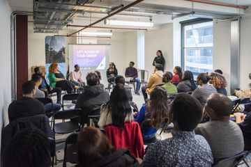 A diverse group of people sitting in a circle in a modern room, attentively listening to a speaker near a screen displaying "Lift - Power Up Your Career."