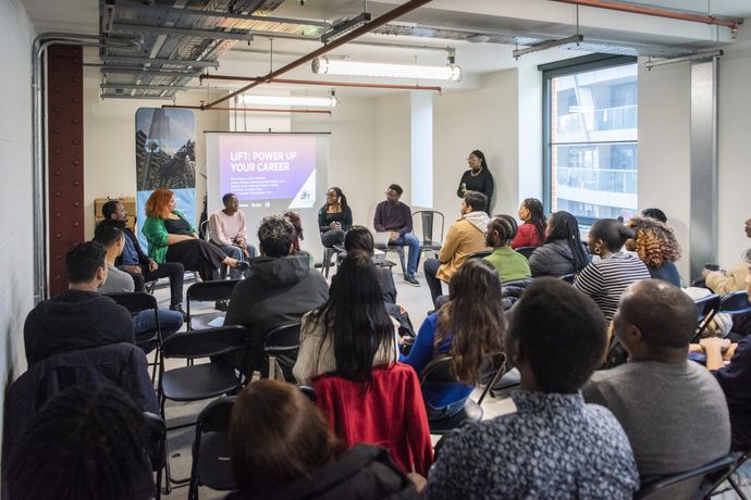 A diverse group of people sitting in a circle in a modern room, attentively listening to a speaker near a screen displaying "Lift - Power Up Your Career."