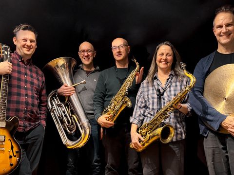 Five musicians smiling, holding instruments: a guitar, tuba, two saxophones, and a cymbal, standing against a dark background.