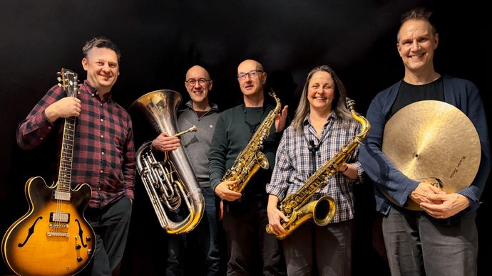 Five musicians smiling, holding instruments: a guitar, tuba, two saxophones, and a cymbal, standing against a dark background.