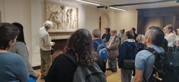 A group of people attentively listening to a guide in a museum, observing a detailed wall sculpture.