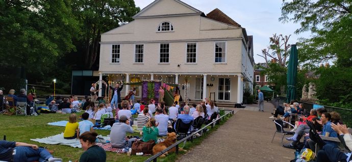 An outdoor crowd watches a small performance in front of a building on a grassy lawn.