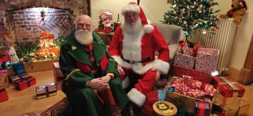 Two men dressed as Santa Claus sit in a festive room with a decorated tree, fireplace, and numerous wrapped gifts around them.