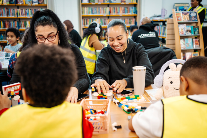 people smile while building lego blocks, children wearing high vis jackets sit with backs to camera playing. people can be seen in background in what appears to be a libary with multiple bookcases behind