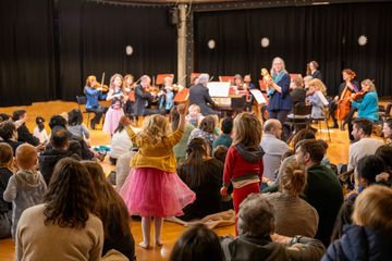 Children sitting on floor with parents, one small girl raises hands in air as orchestra play in background