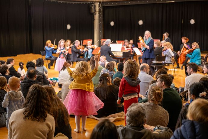 Children sitting on floor with parents, one small girl raises hands in air as orchestra play in background