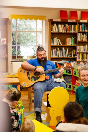Man playing guitar in a library, surrounded by children and parents. Shelves filled with books are visible in the background.