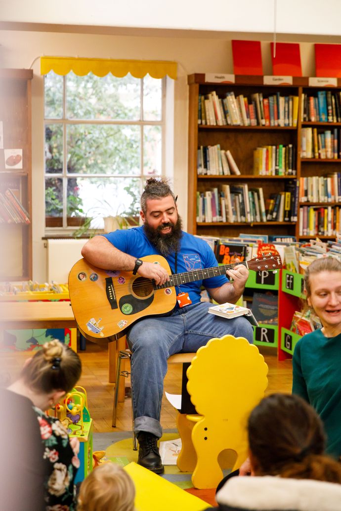 Man playing guitar in a library, surrounded by children and parents. Shelves filled with books are visible in the background.