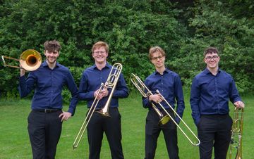 Four men in matching blue shirts stand on grass, each holding a trombone, with trees in the background.