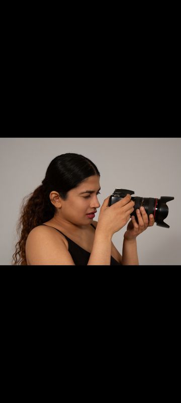 A woman with long curly hair is holding a camera to her eye, focusing on her subject. She is wearing a sleeveless black top.