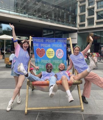 Four people in colorful outfits joyfully pose on a large deck chair at a Pride event, with a vibrant Pride-themed backdrop behind them.