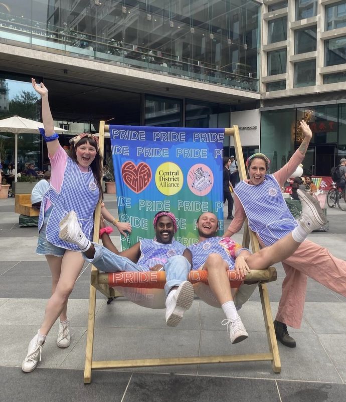 Four people in colorful outfits joyfully pose on a large deck chair at a Pride event, with a vibrant Pride-themed backdrop behind them.