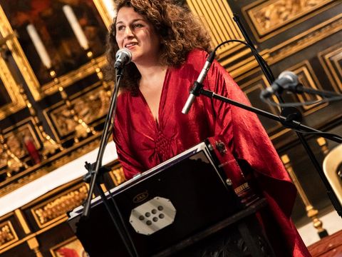in a red robe sings beside microphones and instrument within an ornate venue.
