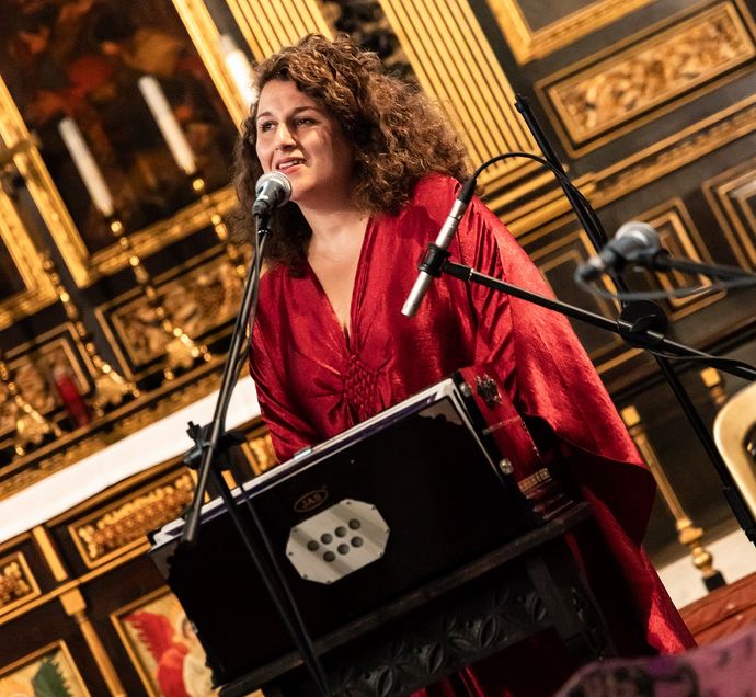 in a red robe sings beside microphones and instrument within an ornate venue.