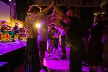 People dancing in a dimly lit room, forming an arch with their arms. A band plays on stage, and purple lighting adds ambiance.