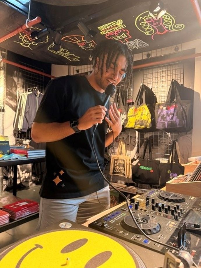 Man with microphone in studio with tote bags behind, in front of decks
