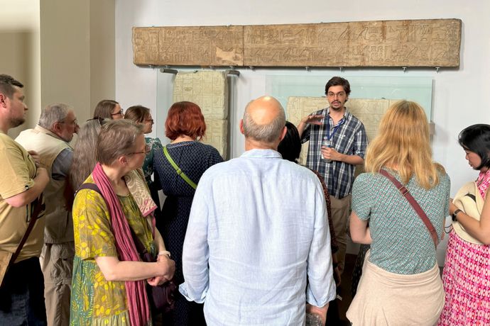 A group of people listens to a guide explaining ancient artifacts displayed on a wall in a museum setting.