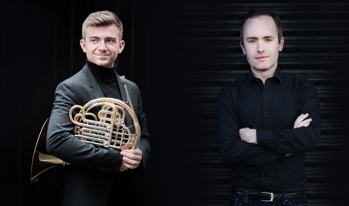 Two men in black attire; one holding a French horn, the other with arms crossed, stand against a dark background.