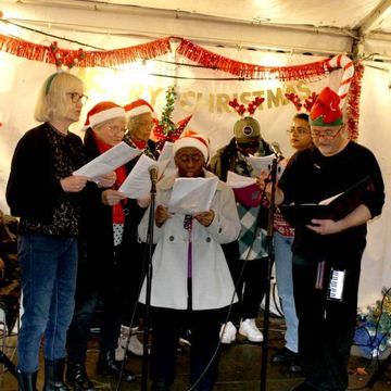 A group of people wearing festive hats sing from sheets in front of microphones, with Christmas decorations in the background.