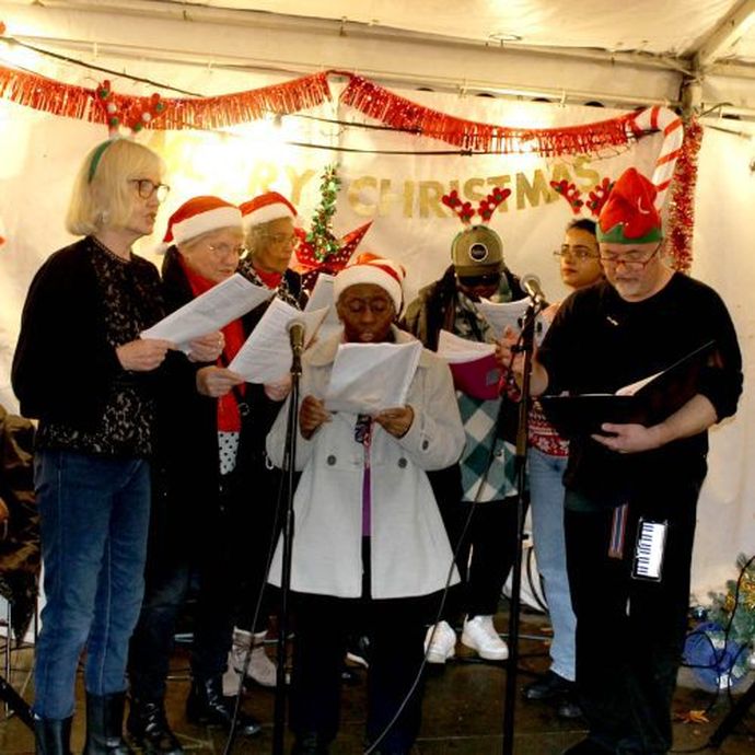 A group of people wearing festive hats sing from sheets in front of microphones, with Christmas decorations in the background.