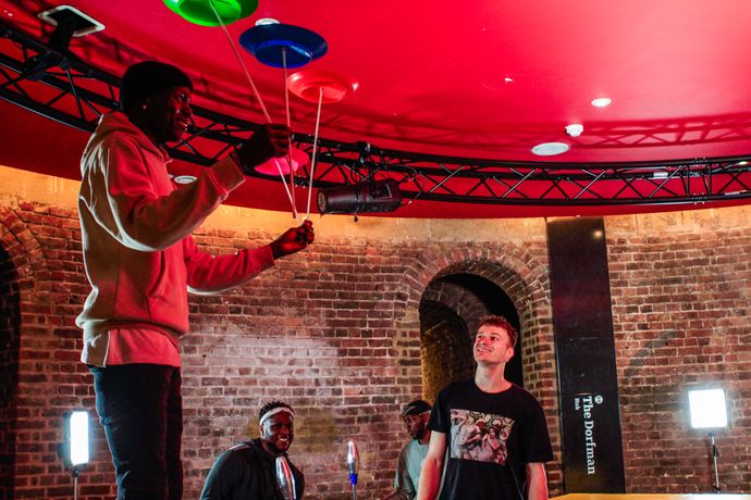 A man balances three plates on sticks as three others look on in a red brick building with studio lights