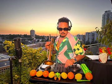 DJ mixing music with headphones on a rooftop at sunset, holding a ladle. Table decorated with fruits and plants.