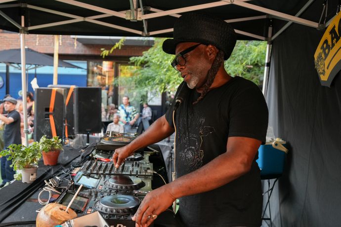 DJ performing outdoors on a turntable, wearing sunglasses and a hat, with plants nearby; audience visible in the background.