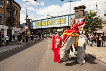 Decorated elephant puppet and performers in vibrant red costumes parade beneath Camden Lock bridge.