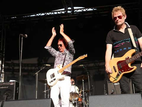 Musicians on stage with one person clapping and another holding a bass guitar, performing under stage lights.