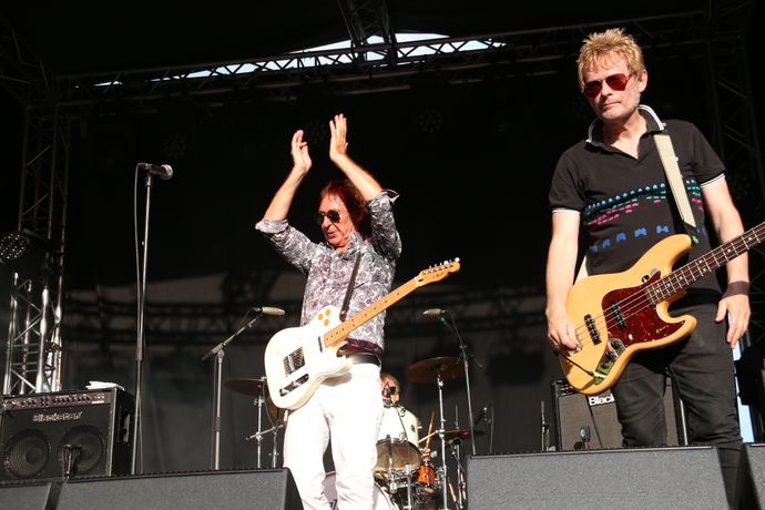 Musicians on stage with one person clapping and another holding a bass guitar, performing under stage lights.