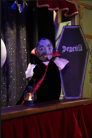 A puppet resembling Dracula stands beside a coffin labeled "Dracula," with a lantern on the table and a starry backdrop.
