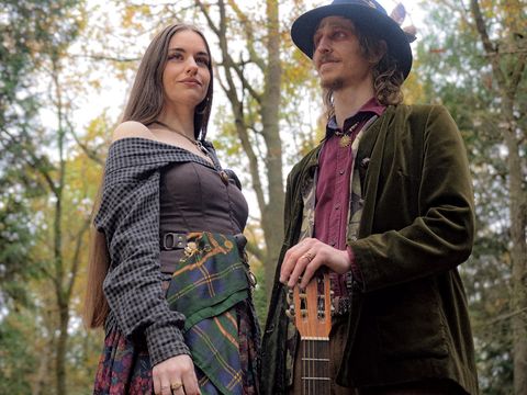 A man and woman in vintage-style clothing stand in a wooded area, the man holding a stringed instrument.