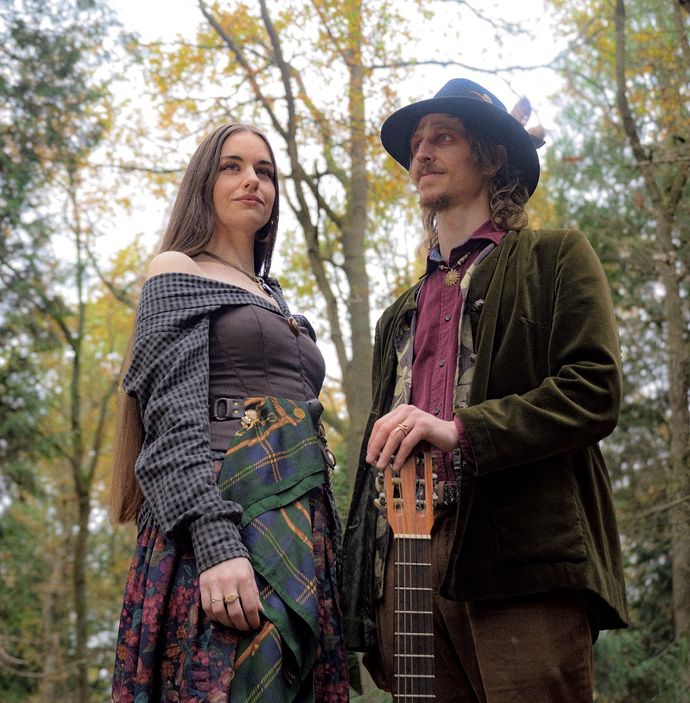 A man and woman in vintage-style clothing stand in a wooded area, the man holding a stringed instrument.
