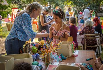 People engaged in arts and crafts at an outdoor community event, surrounded by colorful materials and greenery.