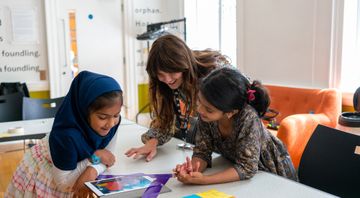 Two young girls and a woman happily engage with a tablet at a table in a bright room.