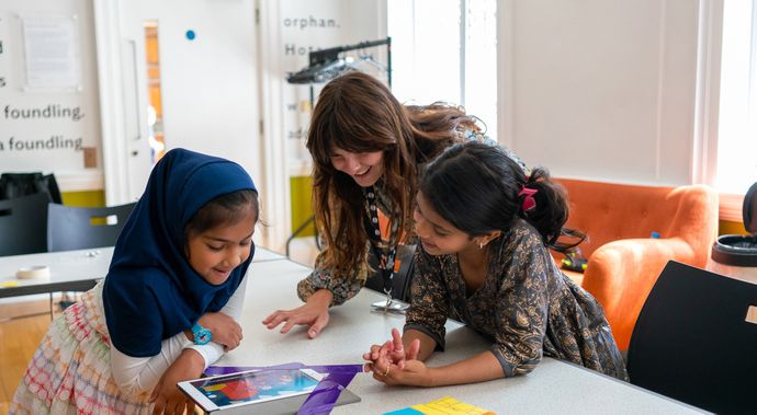 Two young girls and a woman happily engage with a tablet at a table in a bright room.