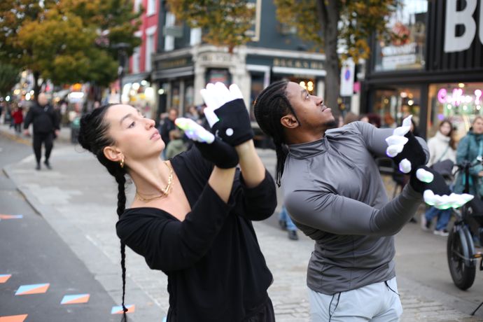 Two people perform a dance routine on a city street, wearing black and gray outfits with glowing gloves, surrounded by onlookers and shops.