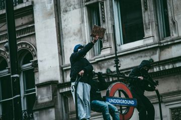 Protesters demonstrate atop a London Underground entrance holding a "Black Lives Matter" sign, against an ornate building backdrop.