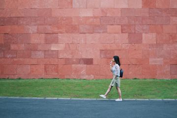 A person walks across a grassy area beside a large wall made of reddish stone blocks.