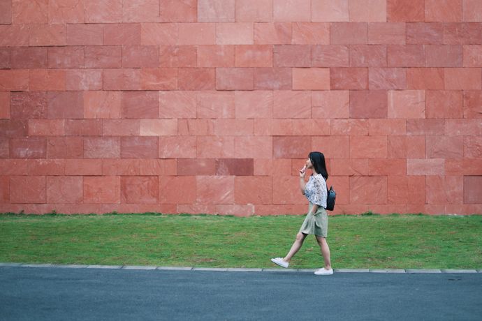 A person walks across a grassy area beside a large wall made of reddish stone blocks.