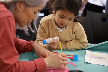 Woman helping a young girl with a craft project, using glue and paper cutouts at a table.