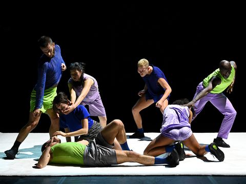 seven people wearing variations of blue, lime green and lilac tops and trouser combos move in different posed shapes