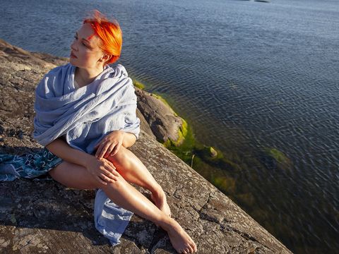 A person with orange hair sits wrapped in a light blue shawl on a rocky shore, looking out over a calm body of water on a sunny day.